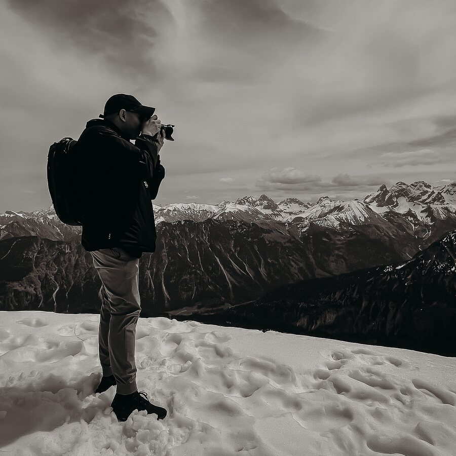 Sam Atagana photographing from a snow-covered ridge in the Swiss Alps, camera raised, pack on his back.